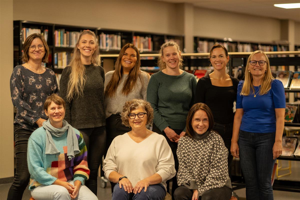 Ni damer står oppstilt i to rekker. Seks stående bak tre som sitter på puffer. Foto. - Klikk for stort bilde