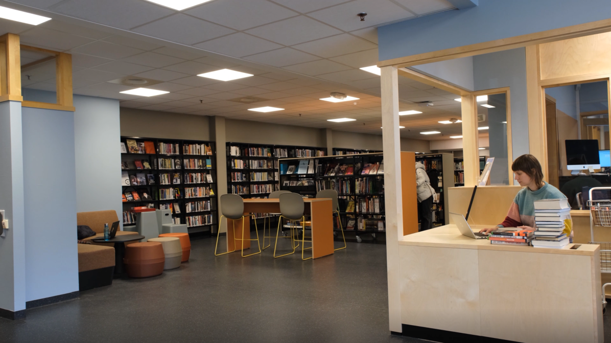 Et biblioteksrom. Til høyre en skranke hvor det sitter en kvinnelig bibliotekar foran en bærbar pc. Foran henne er også to stabler med bøker. Midt i bildet er det et høyt bord med fire stoler. Til venstre en hjørnesofa med bord og puffer. Langs veggen i bakgrunnen er det bokhyller. Det er også bokhyller som står ute i rommet bak bordet. Foto. - Klikk for stort bilde